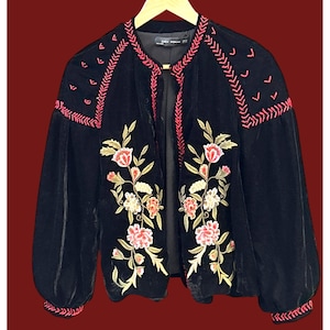 May include: Black velvet jacket with floral embroidery in pink, white, and gold. The jacket features long, puffy sleeves and pink embroidered accents along the collar and cuffs. The jacket is open in the front.