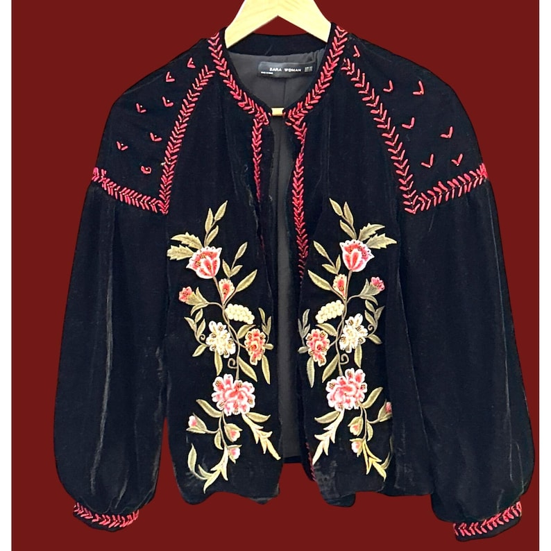 May include: Black velvet jacket with floral embroidery in pink, white, and gold. The jacket features long, puffy sleeves and pink embroidered accents along the collar and cuffs. The jacket is open in the front.