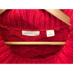 May include: Close-up of a vibrant red knit sweater with a high, rolled neckline. The sweater is hanging on a wooden hanger. The label reads "sleeping on snow". The tag indicates the size is medium and made in China.