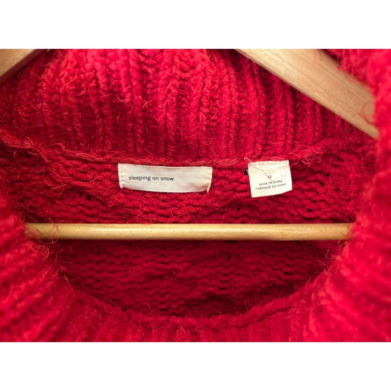 May include: Close-up of a vibrant red knit sweater with a high, rolled neckline. The sweater is hanging on a wooden hanger. The label reads "sleeping on snow". The tag indicates the size is medium and made in China.