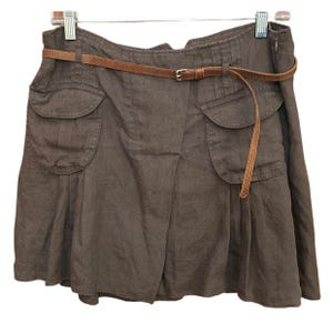 Op de afbeelding: Bruine rok met twee voorzakken en een bruine leren riem. De rok heeft een geplooid ontwerp en hangt aan een metalen hanger. De riem heeft een goudkleurige gesp.