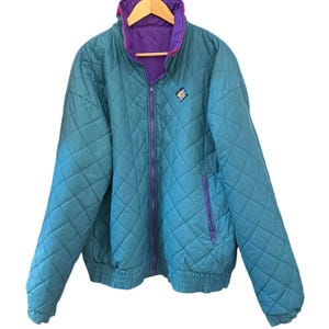 Peut inclure: Une veste matelassée bleu sarcelle avec un motif en losanges, une fermeture éclair violette et un col intérieur violet. La veste présente un petit écusson en forme de losange sur la poitrine gauche avec du texte bleu, jaune et blanc.