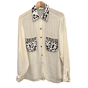 May include: Cream-colored button-down shirt with long sleeves, a collar, and two front pockets. The collar and pockets are embellished with a black and white animal print design. The shirt is hanging on a wooden hanger.