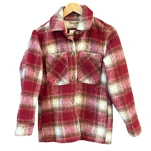 Pode incluir: Casaco xadrez vermelho, branco e castanho com dois bolsos frontais e fecho de botões. O casaco está pendurado num cabide de madeira sobre um fundo branco. O casaco tem gola.