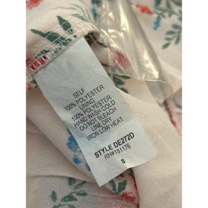 May include: White dress with a floral print and a white tag with care instructions. The tag says "Self 100% Polyester, Lining 100% Polyester, Hand Wash Cold, Do Not Bleach, Line Dry, Iron Low Heat, Style DE272D, RN#151176, S".