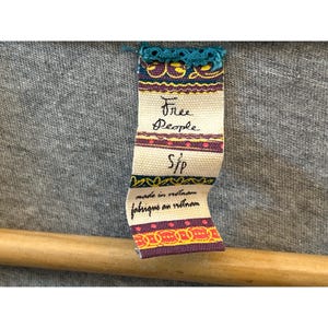 Peut inclure: Gros plan sur un tissu gris avec une &eacute;tiquette attach&eacute;e. L'&eacute;tiquette est de couleur cr&egrave;me avec les mots "Free People" en cursive, et "S/P" en dessous. L'&eacute;tiquette indique &eacute;galement "made in vietnam" et "fabriqu&eacute; en vietnam".