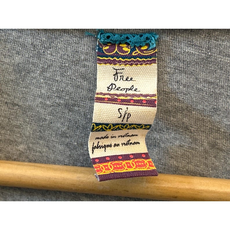 Peut inclure: Gros plan sur un tissu gris avec une &eacute;tiquette attach&eacute;e. L'&eacute;tiquette est de couleur cr&egrave;me avec les mots "Free People" en cursive, et "S/P" en dessous. L'&eacute;tiquette indique &eacute;galement "made in vietnam" et "fabriqu&eacute; en vietnam".