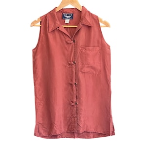 Puede incluir: Una camisa sin mangas de color coral con cuello y un bolsillo. La camisa presenta cierres de palanca decorativos en la parte delantera y se muestra en una percha de madera.