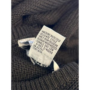 May include: Close-up of a brown knit sweater with a white tag. The tag reads "MACHINE WASH COLD, DELICATE CYCLE, ONLY NON-CHLORINE BLEACH WHEN NEEDED, TUMBLE DRY LOW". The sweater has a textured knit pattern.