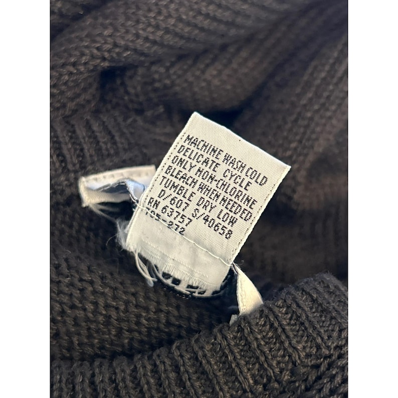 May include: Close-up of a brown knit sweater with a white tag. The tag reads "MACHINE WASH COLD, DELICATE CYCLE, ONLY NON-CHLORINE BLEACH WHEN NEEDED, TUMBLE DRY LOW". The sweater has a textured knit pattern.