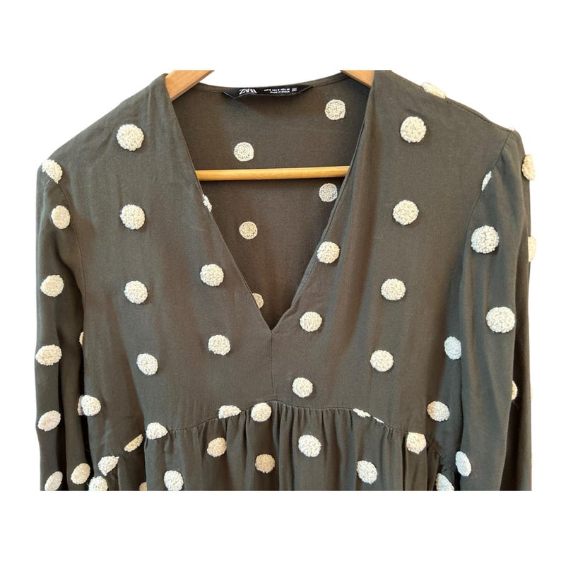 May include: Olive green dress with white, textured polka dots. The dress features a V-neck and long sleeves. The dress is hanging on a wooden hanger.