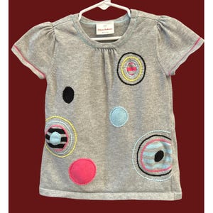 May include: Gray short-sleeved t-shirt with colorful embroidered circles and dots. The shirt has a round neckline and a small tag. The sleeves and neckline are trimmed with pink and light blue.