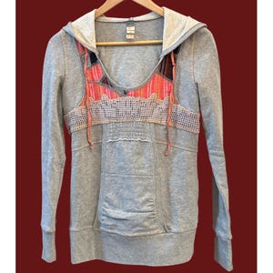 Peut inclure: Sweat &agrave; capuche gris avec un design unique. Le devant pr&eacute;sente un motif patchwork color&eacute; dans les tons de rose, orange et noir, avec une superposition en crochet. Il poss&egrave;de une poche kangourou et des manches longues. Le sweat est suspendu &agrave; un cintre en bois.
