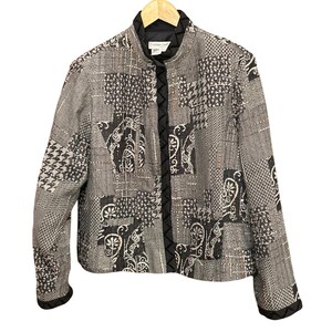 May include: A patchwork jacket in shades of gray, black, and white, featuring various geometric and paisley patterns. The jacket has a stand-up collar, a black trim, and a button closure. The sleeves have a black trim.