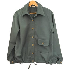 May include: A dark green long-sleeve shirt featuring a pointed collar, a front button-down closure with gold-toned buttons, a single chest pocket with a buttoned flap, and a drawstring hem. The cuffs also feature matching gold-toned buttons.