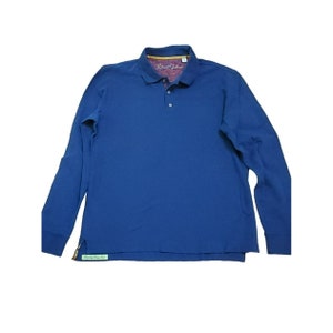 May include: A long-sleeved blue polo shirt with a button-up collar. The shirt has a small green label on the bottom left side that says "Smoking Jacket Club".