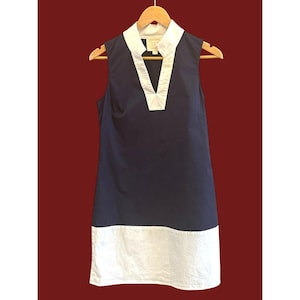 May include: Sleeveless navy blue dress with a white V-neck collar and a white hemline. The dress is hanging on a wooden hanger against a dark red background. The dress has a simple, classic design.