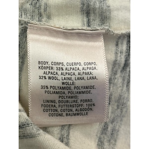 May include: Close-up of a clothing tag with text in multiple languages. The tag lists the fabric composition: 33% alpaca, 32% wool, 35% polyamide, and 100% cotton. The background is a white and gray patterned fabric.