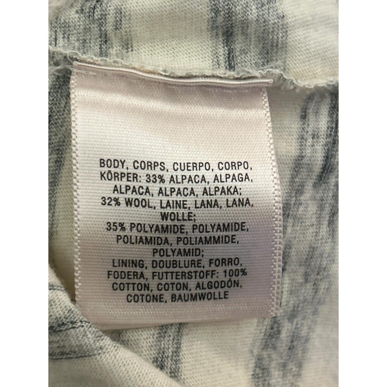 May include: Close-up of a clothing tag with text in multiple languages. The tag lists the fabric composition: 33% alpaca, 32% wool, 35% polyamide, and 100% cotton. The background is a white and gray patterned fabric.