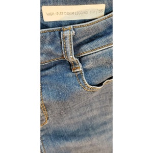 May include: Close-up of a pair of blue denim leggings with a high rise waist. The tag reads "HIGH-RISE DENIM LEGGING Size 26".