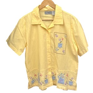 Op de afbeelding: Een gele shirt met korte mouwen, bloemenborduurwerk en knoopsluiting. Het shirt heeft een geborduurde zak en een decoratieve rand met bloempotten, gieters en vlinders. Het label toont "JANE ASHLEY".