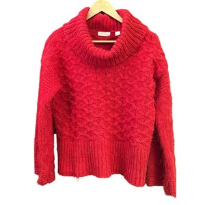 May include: A vibrant red sweater with a cowl neck and long sleeves. The sweater is made of a textured knit fabric and hangs on a wooden hanger. The design features a detailed pattern across the body.
