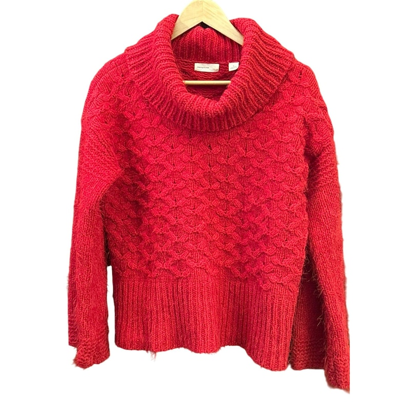 May include: A vibrant red sweater with a cowl neck and long sleeves. The sweater is made of a textured knit fabric and hangs on a wooden hanger. The design features a detailed pattern across the body.