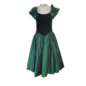 May include: A knee-length dress featuring a dark green velvet bodice with a square neckline and short, ruffled sleeves adorned with fabric rosettes. The skirt is made of a shimmering, iridescent green taffeta fabric with a full, pleated silhouette.