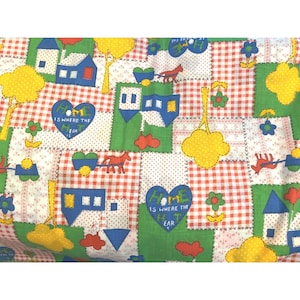 Op de afbeelding: Een patchworkstof met een herhalend patroon van huizen, bomen, harten en paardenwagens. Het ontwerp bevat rood geruite, groene en witte stippen vierkanten. De tekst "Home is where the heart is" is zichtbaar.