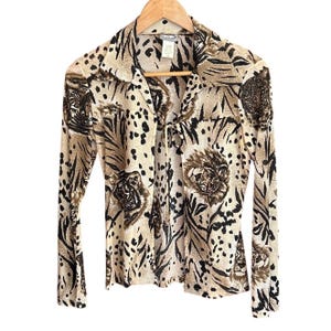 Könnte beinhalten: Eine langärmlige Bluse mit Tigerprint-Design. Die Bluse hat einen Kragen und einen Krawattenverschluss. Der Stoff ist beige mit schwarzen und braunen Tigerstreifen und -flecken. Die Bluse hängt an einem Holzkleiderbügel.
