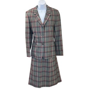 Op de afbeelding: Een grijze geruite rokpak met een blazer en rok. De blazer heeft een reverskraag, twee voorzakken en een sluiting met twee knopen. De rok is een klassieke A-lijn stijl. Het ruitpatroon bevat grijze, rode en zwarte lijnen.