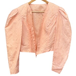 Puede incluir: Una chaqueta corta de color melocotón claro con mangas largas de hombros abullonados fruncidos y un sutil patrón similar a la veta de la madera en toda la tela. La chaqueta tiene un diseño frontal abierto con cuello en V y sin botones.