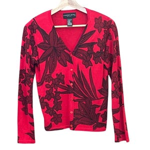 Puede incluir: Cárdigan rojo con cuello en V y mangas largas, adornado con un diseño floral oscuro. La etiqueta dice "Aubrey Hilton". El cárdigan se abrocha con botones. La prenda está colgada en una percha de madera.
