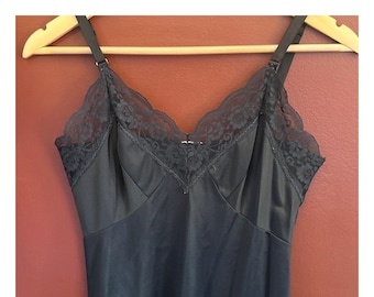Puede incluir: Un vestido lencero negro con tirantes ajustables y escote en V. El escote y los tirantes están adornados con delicado encaje negro. El vestido está colgado de una percha de madera sobre un fondo marrón rojizo.
