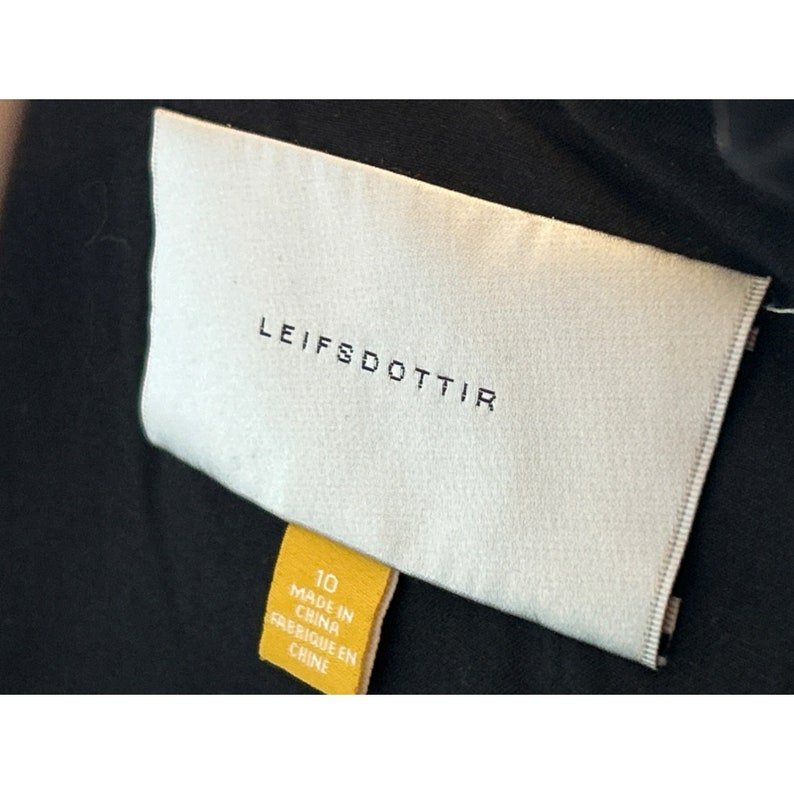 Op de afbeelding: Een wit label met de tekst "LEIFSDOTTIR" in zwarte letters. Een geel label met de tekst "10 MADE IN CHINA FABRIQUE EN CHINE" is bevestigd aan het witte label.