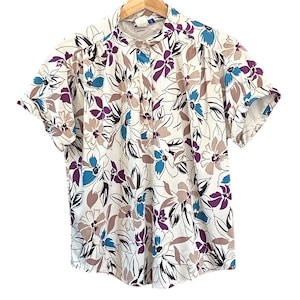 Puede incluir: Una blusa de manga corta con estampado floral. La blusa presenta un fondo blanco con grandes flores en tonos morados, azules y beige. Tiene cuello mandarín y cierre de botones. La blusa está colgada de una percha de madera.