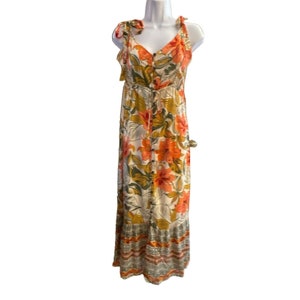 May include: A white maxi dress with a floral print in shades of orange, green, and yellow. The dress has a V-neckline, adjustable straps, and a button-up front. The hemline is tiered and features a geometric pattern in shades of orange, pink, and gray.