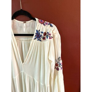 May include: A white, long-sleeved, embroidered peasant top with a V-neckline and bell sleeves. The embroidery features blue, pink, and red flowers.