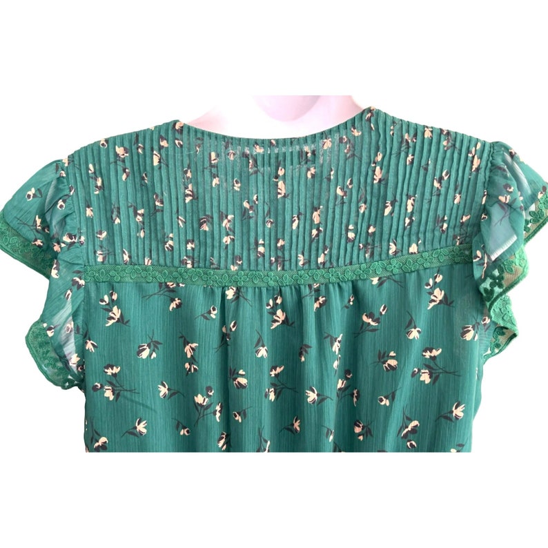 May include: A green floral print blouse with a ruffled neckline and short sleeves. The blouse has a lace trim around the neckline.
