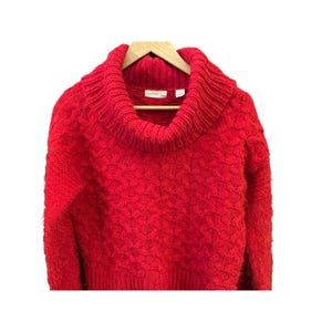 May include: A vibrant red sweater with a chunky knit design and a wide, cozy cowl neck. The sweater is displayed on a wooden hanger against a white background, showcasing its textured pattern and warm color.
