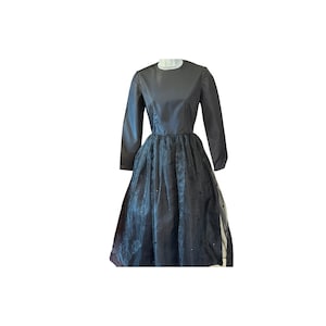 Könnte beinhalten: Ein langärmeliges Kleid mit einem einfarbigen, dunkelgrauen Oberteil mit Rundhalsausschnitt und einem voluminösen, transparenten dunkelgrauen Rocküberwurf. Der Rock ist mit kleinen, gleichmäßig verteilten dunklen Akzenten verziert.