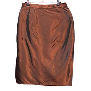 Op de afbeelding: Een knielange rok in een effen, iriserende koperbruine kleur. Het kledingstuk heeft een gladde, licht reflecterende afwerking, een gestructureerde tailleband en een recht model dat verticaal aan een metalen kledinghanger hangt.