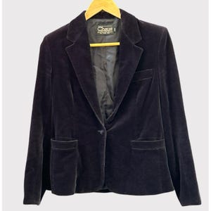 Op de afbeelding: Een donker marineblauwe fluwelen blazer met een enkele knoopsluiting, twee voorzakken en een revers met inkeping. De blazer hangt aan een houten kleerhanger. De merknaam "Chaus" is zichtbaar op het label.