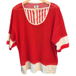 May include: A red mesh short-sleeve shirt featuring a white and red vertical striped henley-style inset at the neckline with three buttons. The shirt has a wide white band at the bottom hem and white bands on the sleeves. A small label is at the neck.