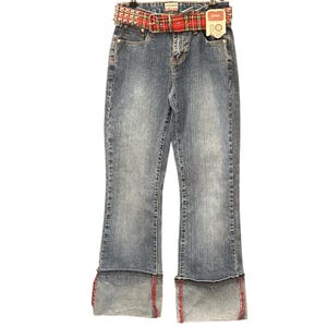 May include: Blue denim jeans with a red plaid belt and a studded buckle. The jeans have a flared leg and a folded cuff with red stitching. A tag is attached to the jeans.