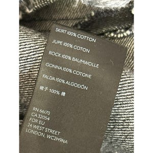 May include: A black fabric label with white text detailing the composition of a skirt in multiple languages. The text reads "SKIRT 100% COTTON, JUPE 100% COTON, ROCK 100% BAUMWOLLE, GONNA 100% COTONE, FALDA 100% ALGOD&Oacute;N, 裙子 100%棉". The label also includes the company's contact information: "RN 66170, CA 32054, FOR EU, 24 WEST STREET, LONDON, WC2H9NA".