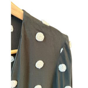 May include: Close-up of a dark green garment with large, textured, off-white polka dots. The fabric is draped over a wooden hanger. The garment has a V-neckline and appears to be a top or dress.