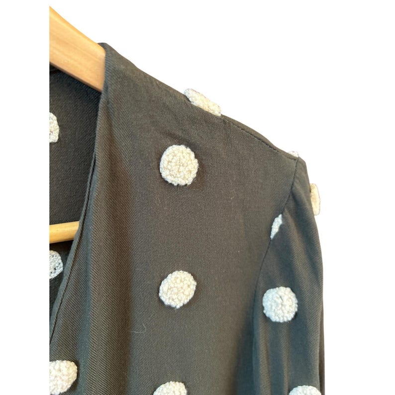 May include: Close-up of a dark green garment with large, textured, off-white polka dots. The fabric is draped over a wooden hanger. The garment has a V-neckline and appears to be a top or dress.