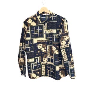 May include: A long-sleeved button-down shirt featuring a patchwork pattern in navy blue, cream, and dark green. The design consists of various geometric squares, floral motifs, and circular accents arranged in a grid-like layout on the fabric.