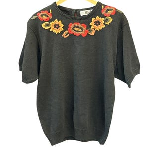 May include: Dark gray short-sleeve sweater with embroidered floral design in red, yellow, and green. The crew neck sweater has a keyhole back and is hanging on a wooden hanger. The word "SEGU" is visible on the back of the sweater.
